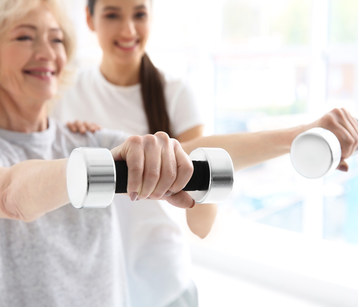 Frau trainiert mit zwei Hanteln, während eine Trainerin sie unterstützt